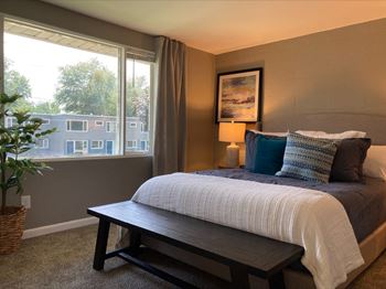 A bedroom with a bed, a bench, and a window overlooking a building at Townhomes at 28th, Columbus, IN, 47201